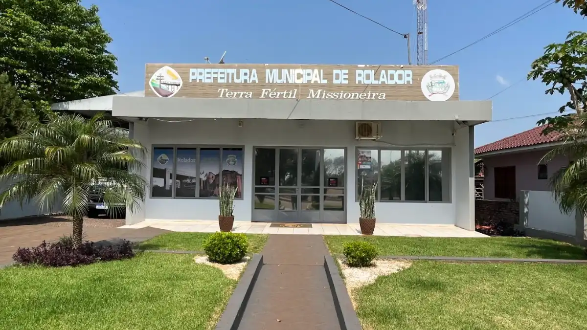 Fachada da Prefeitura Municipal de Rolador, no Rio Grande do Sul. O prédio térreo cinza possui gramado frontal, palmeiras laterais e um letreiro com a frase Terra Fértil Missioneira sob o céu azul.