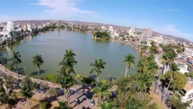 Vista aérea da Lagoa Paulino em Sete Lagoas/MG, com palmeiras ao redor, vias urbanas e prédios ao fundo em dia ensolarado. Foto: Luiz Claudio Alvarenga/Wikipédia