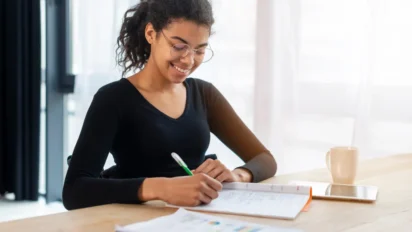 Mulher jovem usando óculos, sorrindo enquanto escreve em um caderno sobre uma mesa, em ambiente iluminado, com caneca e celular ao lado, representando estudo ou preparação para concurso.
