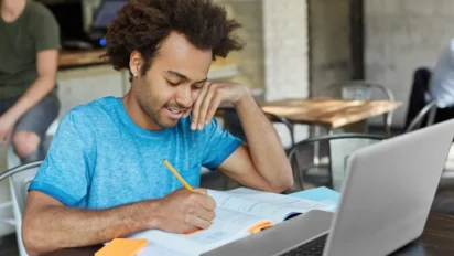 Jovem estudando em uma mesa com caderno e laptop, escrevendo com lápis enquanto consulta anotações em um ambiente interno tipo café ou biblioteca. Foto: wayhomestudio/Freepik