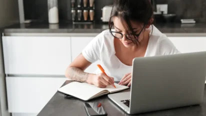Jovem mulher estudando em casa, escrevendo em um caderno enquanto usa um notebook sobre a mesa. Foto: drobotdean/Freepik