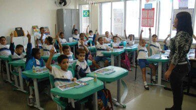Processos Seletivos 8 Sala de aula com alunos uniformizados sentados em carteiras verdes, muitos com a mão levantada, e professora em pé à frente da turma. Foto: Divulgação/Prefeitura do Rio de Janeiro-RJ