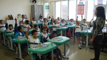 Sala de aula com alunos uniformizados sentados em carteiras verdes, muitos com a mão levantada, e professora em pé à frente da turma. Foto: Divulgação/Prefeitura do Rio de Janeiro-RJ