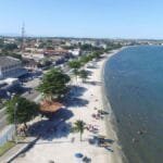Vista aérea da orla de Iguaba Grande, com faixa de areia branca, mar calmo e diversas pessoas na praia e no mar; ao lado, uma avenida com comércios e residências.
