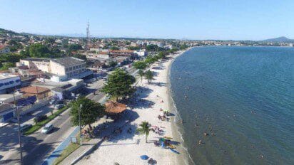 Vista aérea da orla de Iguaba Grande, com faixa de areia branca, mar calmo e diversas pessoas na praia e no mar; ao lado, uma avenida com comércios e residências.