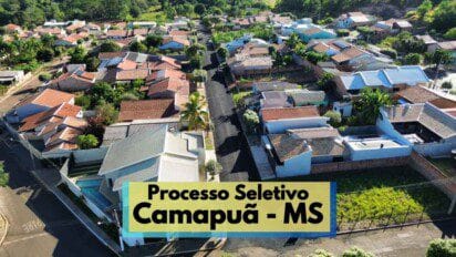 Imagem aérea de um bairro residencial em Camapuã, no Mato Grosso do Sul, mostrando ruas asfaltadas e casas com telhados variados cercadas por vegetação. Ao centro, destaca-se uma rua principal alinhada com árvores. No canto inferior, há uma faixa com os dizeres “Processo Seletivo Camapuã - MS”, indicando a realização de um processo seletivo na cidade, com destaque visual chamativo.