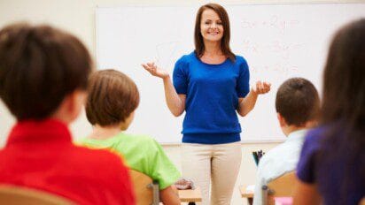 Professora sorridente em sala de aula explicando conteúdo a alunos sentados, com quadro branco ao fundo contendo equações matemáticas.