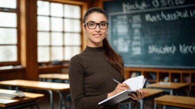 Processos Seletivos 4 Mulher com óculos segura caderno e caneta em sala de aula com carteiras e quadro negro ao fundo.