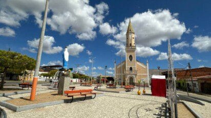 Praça urbana em Palhano (CE) com estátua de Nossa Senhora, fonte central, bancos vermelhos, igreja ao fundo, calçamento em pedras claras e céu azul com nuvens.