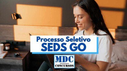 Mulher jovem com cabelos castanhos longos e lisos, vestindo camiseta branca, está sentada em um quarto com decoração moderna, sorrindo enquanto utiliza um notebook no colo; ao centro da imagem há uma sobreposição gráfica com os dizeres "Processo Seletivo SEDS GO" em letras azuis e "MDC Concursos" em destaque na parte inferior da arte