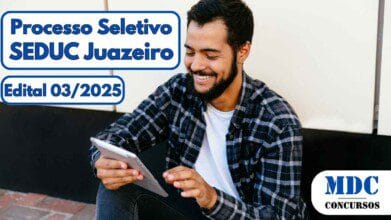 Homem jovem com barba e camisa xadrez sentado no chão, sorrindo enquanto usa um tablet em ambiente externo com parede clara ao fundo - Processo Seletivo SEDUC Juazeiro (BA) oferece salários de até R$ 7,7 mil - Edital 03/2025