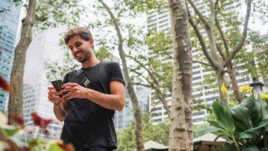 Processos Seletivos 2 Homem sorridente usando celular em parque urbano com prédios ao fundo e árvores ao redor. Foto: Freepik