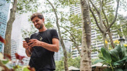 Homem sorridente usando celular em parque urbano com prédios ao fundo e árvores ao redor. Foto: Freepik