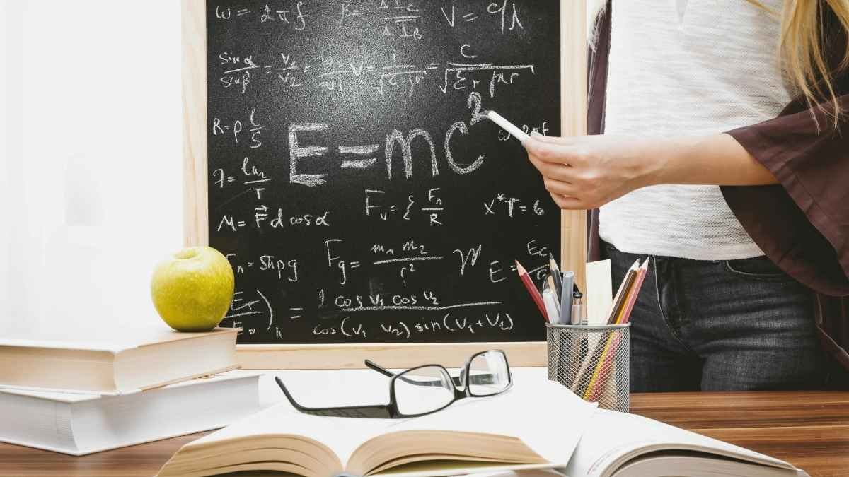Pessoa diante de quadro-negro com fórmulas de física, segurando giz; sobre a mesa, livros, maçã verde, óculos e porta-lápis com materiais escolares.