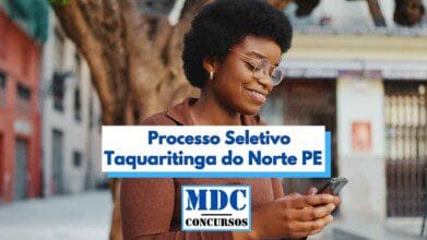 Mulher negra de cabelos curtos e cacheados, usando óculos e camisa marrom, sorri enquanto olha para o celular. Ao fundo, há uma rua com prédios e uma grande árvore. Sobre a imagem, há uma faixa com os dizeres “Processo Seletivo Taquaritinga do Norte PE” e, abaixo, o logotipo do site MDC Concursos em azul e preto.
