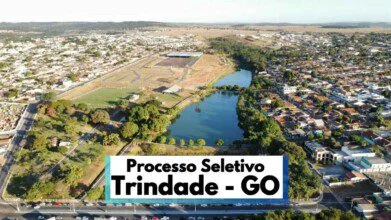 A imagem mostra uma vista aérea panorâmica da cidade de Trindade, no estado de Goiás, destacando áreas urbanas com casas e prédios, além de um grande lago cercado por vegetação e vias asfaltadas. No canto inferior da imagem, há uma faixa com os dizeres "Processo Seletivo Trindade - GO" em letras grandes e pretas. O cenário urbano se estende ao fundo, com uma mescla de áreas verdes e construções residenciais e comerciais. A imagem transmite a dimensão e organização da cidade, com destaque para a estrutura urbana e a presença de áreas de lazer e natureza.