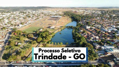 A imagem mostra uma vista aérea panorâmica da cidade de Trindade, no estado de Goiás, destacando áreas urbanas com casas e prédios, além de um grande lago cercado por vegetação e vias asfaltadas. No canto inferior da imagem, há uma faixa com os dizeres "Processo Seletivo Trindade - GO" em letras grandes e pretas. O cenário urbano se estende ao fundo, com uma mescla de áreas verdes e construções residenciais e comerciais. A imagem transmite a dimensão e organização da cidade, com destaque para a estrutura urbana e a presença de áreas de lazer e natureza.