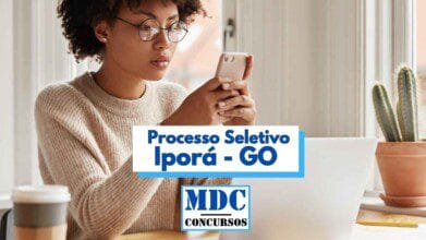 Mulher jovem com cabelos cacheados e óculos está sentada em frente a uma janela, usando um celular com atenção. Ela veste um suéter bege e está ao lado de um notebook branco e um copo térmico. Ao fundo, há vasos com cactos em uma mesa de madeira. Sobre a imagem, há um banner escrito “Processo Seletivo Iporá - GO” e o logotipo “MDC Concursos”.