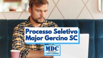 Homem jovem de barba curta e cabelo castanho claro usando camisa xadrez amarela e preta, concentrado enquanto utiliza um notebook branco em um ambiente interno com parede decorada ao fundo e sofá azul-escuro, com um copo descartável rosa sobre a mesa; sobre a imagem há uma faixa branca com a inscrição Processo Seletivo Major Gercino SC em letras azuis e abaixo o logotipo MDC Concursos