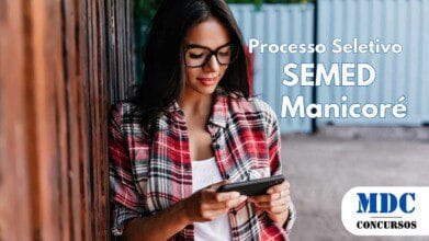 Processos Seletivos 8 Mulher jovem de cabelos escuros e longos, usando óculos de grau e camisa xadrez vermelha sobre uma blusa branca, encostada em uma cerca de madeira enquanto mexe no celular com um leve sorriso. O fundo mostra contêineres azuis e vegetação - Processo Seletivo em Manicoré (AM) abre 100 vagas para Agente de Educação