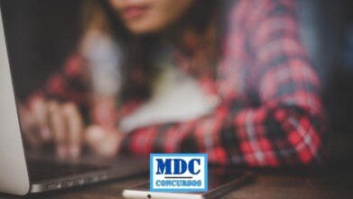 Mulher usando uma camisa xadrez vermelha e preta aparece desfocada ao fundo enquanto digita em um notebook em uma mesa de madeira; em primeiro plano está um smartphone e parte do notebook, ambos em foco; ao centro inferior da imagem há um logotipo com fundo branco e bordas azuis contendo as palavras MDC Concursos em letras azuis - Processo Seletivo em Mariano Moro RS