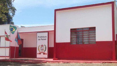 Fachada do prédio da Prefeitura de Painel, localizado no estado de Santa Catarina. A construção é térrea, com pintura em vermelho e branco. À frente do edifício, estão hasteadas quatro bandeiras: a do Brasil, a de Santa Catarina e outras duas bandeiras locais. Na entrada, há uma placa com o brasão do município e os dizeres “Prefeitura de Painel”. O céu está limpo e azul.