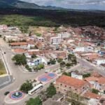 Imagem aérea da cidade de Riacho das Almas, em Pernambuco, mostrando uma vista panorâmica com destaque para a igreja matriz de arquitetura histórica no canto esquerdo inferior. Ao redor, há ruas pavimentadas, casas com telhados de barro, construções residenciais e comerciais. Também é visível uma pequena praça com quadra esportiva e vegetação ao fundo, além de uma área de mata fechada nas proximidades da zona urbana.