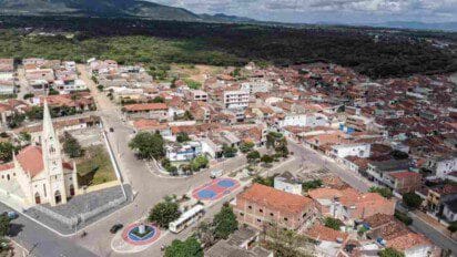 Imagem aérea da cidade de Riacho das Almas, em Pernambuco, mostrando uma vista panorâmica com destaque para a igreja matriz de arquitetura histórica no canto esquerdo inferior. Ao redor, há ruas pavimentadas, casas com telhados de barro, construções residenciais e comerciais. Também é visível uma pequena praça com quadra esportiva e vegetação ao fundo, além de uma área de mata fechada nas proximidades da zona urbana.