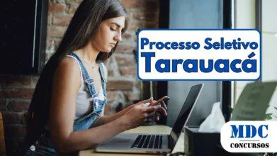 Jovem de cabelos longos e castanhos, vestindo jardineira jeans e blusa branca, sentada em um café com parede de tijolos. Ela segura um celular e tem um laptop aberto à sua frente sobre uma mesa de madeira, com óculos e uma caixa de lenços ao lado. Há uma janela grande à direita com luz natural - Processo Seletivo em Tarauacá (AC) Abre 47 Vagas na Educação