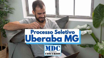 Homem com barba e cabelo curto, vestindo camiseta cinza, está sentado confortavelmente em uma poltrona, sorrindo enquanto utiliza um notebook. Ao fundo, há uma janela e plantas decorativas. Em destaque na imagem, aparece um banner com os dizeres “Processo Seletivo Uberaba MG” e o logotipo “MDC Concursos”, sugerindo que ele está acessando informações sobre concursos públicos na cidade de Uberaba, Minas Gerais.