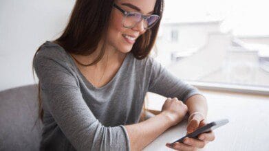 Processos Seletivos 5 Mulher sorridente com óculos e blusa cinza olhando para o celular sentada à mesa próxima a uma janela.