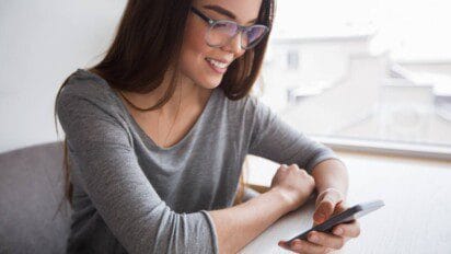 Mulher sorridente com óculos e blusa cinza olhando para o celular sentada à mesa próxima a uma janela.