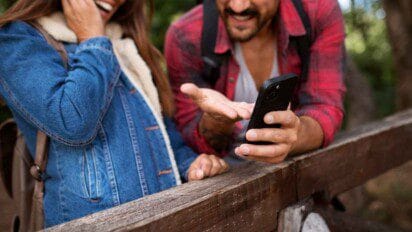 Duas pessoas com mochilas interagem sorrindo com um celular em área ao ar livre; vestem roupas casuais e estão apoiadas em cerca, com vegetação ao fundo.