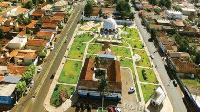 Vista aérea de praça central em Joviânia-GO, com jardins, edifícios públicos e cúpula branca ao fundo, rodeada por ruas e casas.