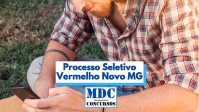 Homem com camisa xadrez colorida e barba ruiva utiliza um celular enquanto está sentado ao ar livre, sobre uma superfície de grama. No centro da imagem, há um destaque com a frase “Processo Seletivo Vermelho Novo MG” em letras azuis, acompanhada do logotipo “MDC Concursos”. A imagem ilustra o uso da internet para inscrições em concursos públicos.