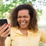 A imagem mostra uma mulher sorridente e visivelmente feliz enquanto segura um celular em uma das mãos. Ela está ao ar livre, em um ambiente com árvores e grama, sugerindo um parque. Veste uma camisa amarela de manga curta e esmalte vermelho nas unhas. A mulher aparenta estar comemorando alguma boa notícia recebida pelo celular, com o braço direito levantado em gesto de comemoração e expressão de entusiasmo no rosto.