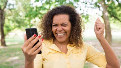 Processos Seletivos 5 A imagem mostra uma mulher sorridente e visivelmente feliz enquanto segura um celular em uma das mãos. Ela está ao ar livre, em um ambiente com árvores e grama, sugerindo um parque. Veste uma camisa amarela de manga curta e esmalte vermelho nas unhas. A mulher aparenta estar comemorando alguma boa notícia recebida pelo celular, com o braço direito levantado em gesto de comemoração e expressão de entusiasmo no rosto.