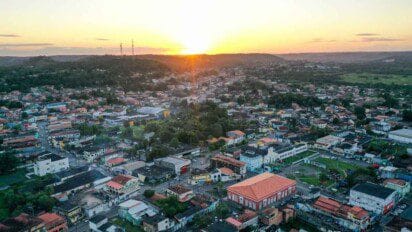 Vista aérea de uma cidade ao entardecer, com muitas casas, prédios baixos e áreas arborizadas, cercada por morros ao fundo e o sol se pondo no horizonte.