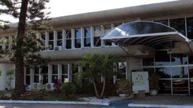 Processos Seletivos 7 Fachada da sede da Seplan na Bahia, com grandes janelas de vidro, cobertura curva na entrada e área externa com árvores e plantas ornamentais.