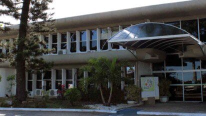 Fachada da sede da Seplan na Bahia, com grandes janelas de vidro, cobertura curva na entrada e área externa com árvores e plantas ornamentais.