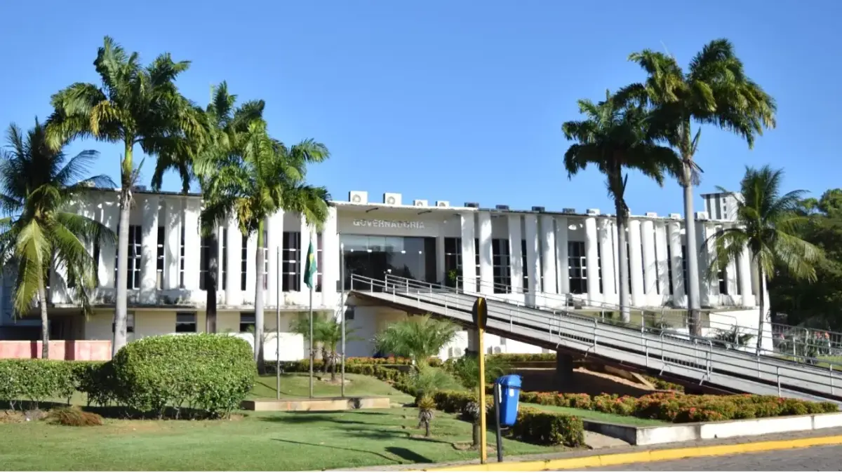 RN abre Concurso Unificado: Fachada do prédio da Governadoria do Rio Grande do Norte em Natal com gramado e palmeiras sob céu azul claro