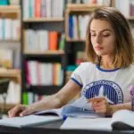 Mulher estudando em uma biblioteca, lendo e fazendo anotações em um caderno. Foto: Freepik