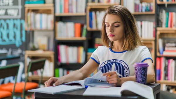 Mulher estudando em uma biblioteca, lendo e fazendo anotações em um caderno. Foto: Freepik