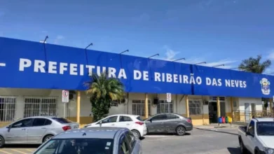 Fachada da Prefeitura de Ribeirão das Neves com placa azul exibindo o nome da instituição, carros estacionados em frente ao prédio e céu azul ao fundo. Foto: Divulgação/Prefeitura de Ribeirão das Neves-MG