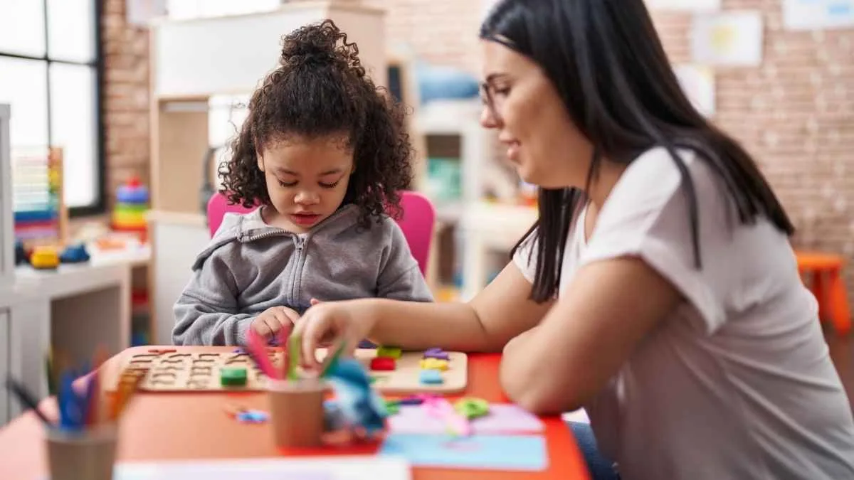Mulher auxilia criança em atividade com letras de madeira em sala de aula infantil colorida, com brinquedos e materiais pedagógicos, destacando ambiente de educação e interação.