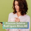 Mulher jovem de cabelos castanhos ondulados, vestindo camisa social rosa claro, olha para um celular em sua mão com expressão de surpresa ou interesse. Ela está em frente a um fundo verde e, sobre a imagem, há um banner com os dizeres “Processo Seletivo Rodrigues Alves AC” em letras brancas sobre fundo verde e azul, indicando conteúdo informativo sobre seleção pública.