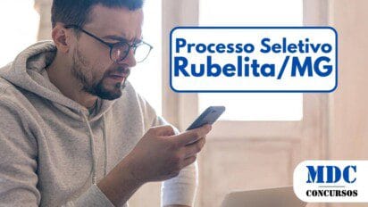 Homem de barba curta e óculos usa um moletom cinza e segura um smartphone com expressão de confusão ou preocupação. Ele está sentado em um ambiente iluminado por luz natural, com um laptop aberto à sua frente e uma janela ao fundo - Rubelita MG oferece vagas com salários de até R$ 3 mil em Processo Seletivo