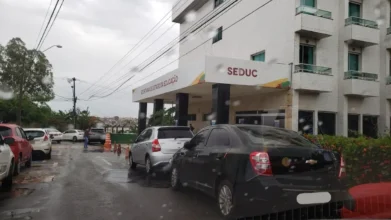 Fachada do prédio da Secretaria de Estado da Educação do Maranhão (SEDUC) em um dia chuvoso, com carros estacionados à frente e identificação na marquise branca.