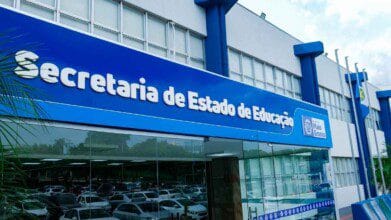 A imagem mostra a fachada da Secretaria de Estado de Educação de Mato Grosso (SEDUC MT). A edificação possui arquitetura moderna, com grandes janelas de vidro e estrutura nas cores azul e branca. Na parte superior, há uma grande placa azul com os dizeres "Secretaria de Estado de Educação" em letras brancas. Ao lado, está o logotipo do Governo de Mato Grosso. Na frente do prédio, há bandeiras hasteadas e um estacionamento com diversos veículos.