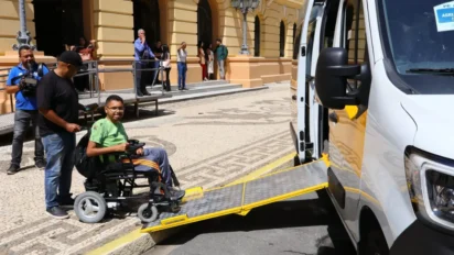Um homem em uma cadeira de rodas utiliza a rampa de acessibilidade de uma van branca e amarela. Ele é auxiliado por outro homem, enquanto outras pessoas observam ao fundo em frente a um prédio histórico. Foto: Divulgação/SEE